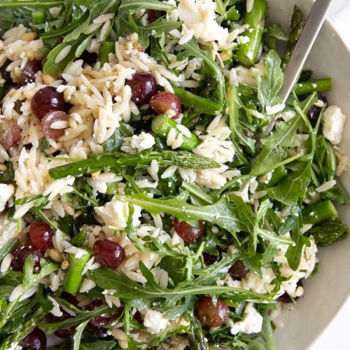 Image of a large salad bowl filled with orzo and arugula salad filled with feta cheese and grapes.