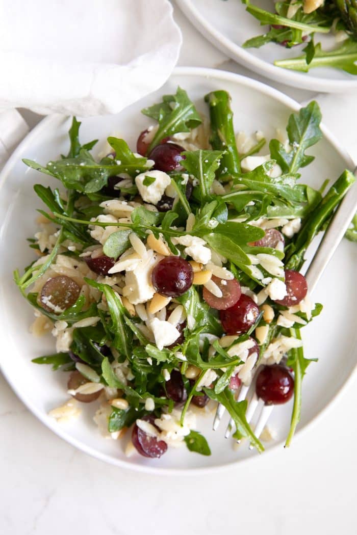 Small white serving plate filled with a salad made with cooked orzo, arugula, asparagus, pine nuts, grapes, and feta cheese.