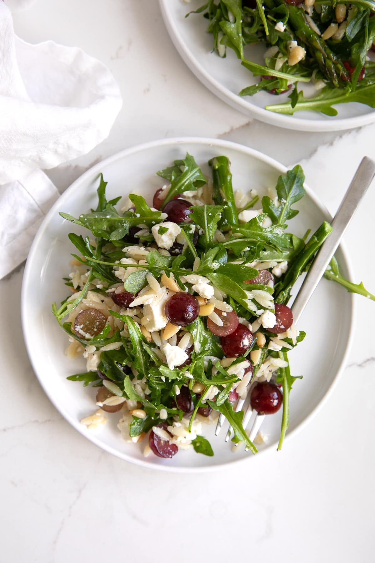 Overhead image of two small white serving plate filled with a salad made with cooked orzo, arugula, asparagus, pine nuts, grapes, and feta cheese.