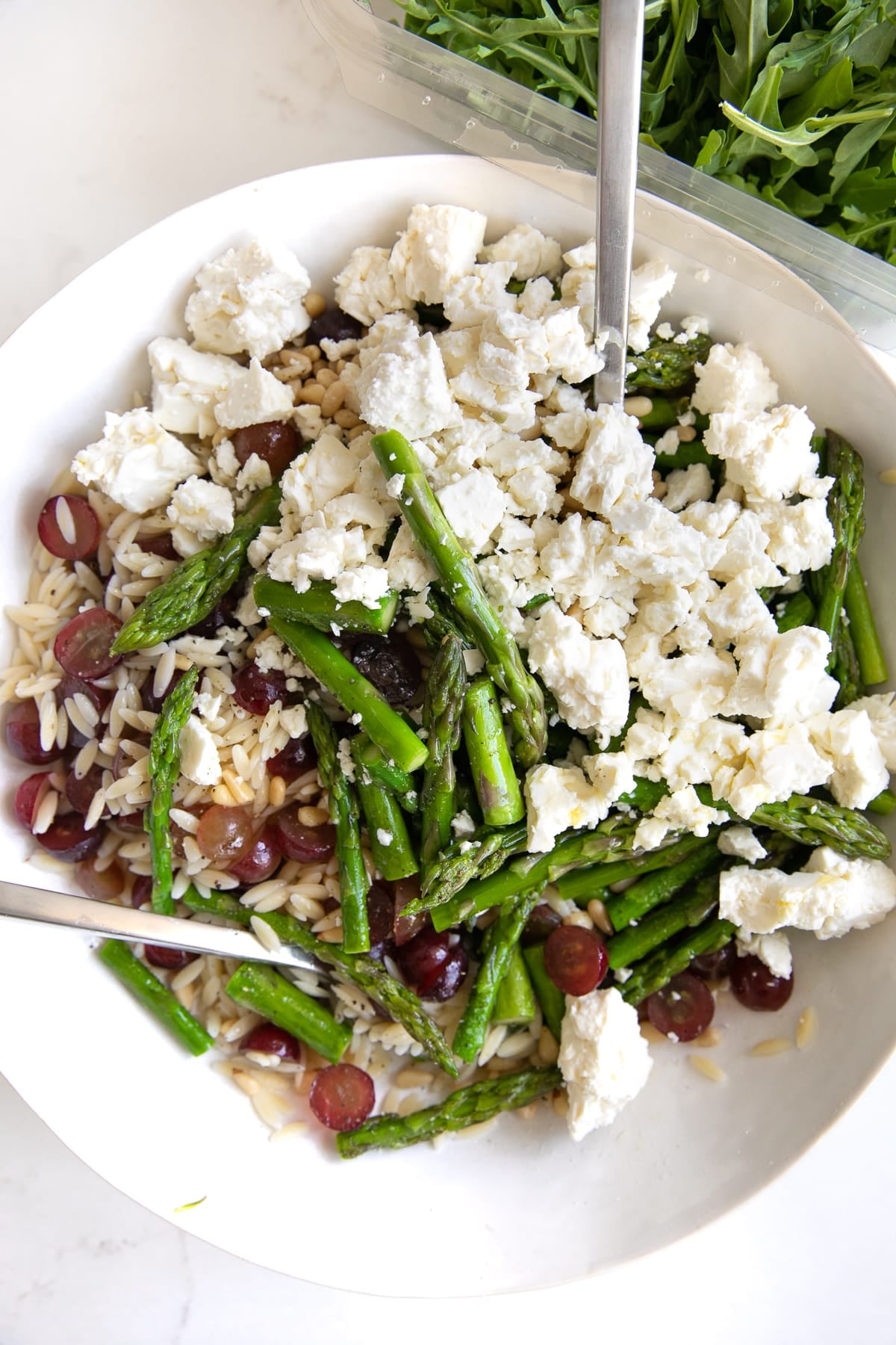 Large salad bowl filled with unmixed cooked orzo, arugula, cooked asparagus, pine nuts, grapes, and feta cheese.