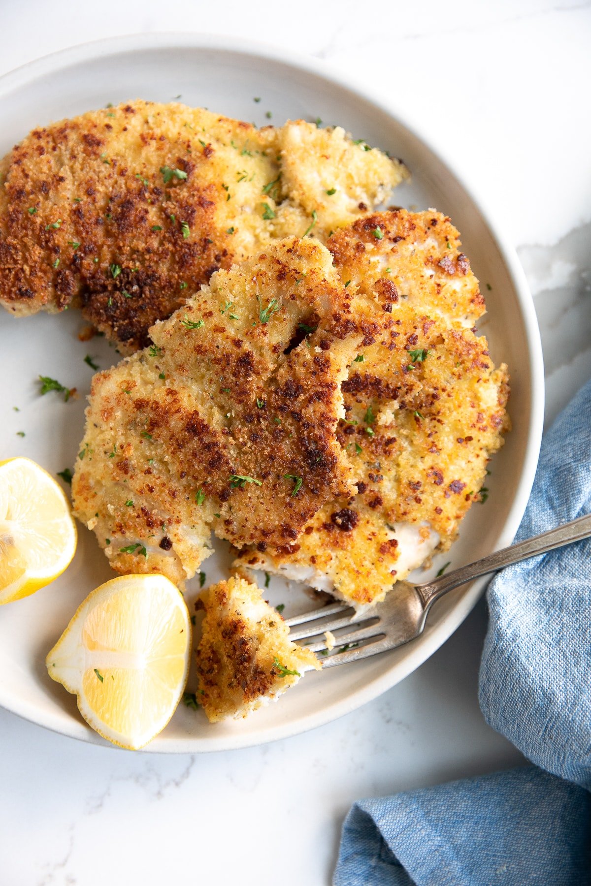 Two thinly sliced parmesan crusted chicken breast pieces on a small white serving plate garnished with chopped parsley and fresh lemon.