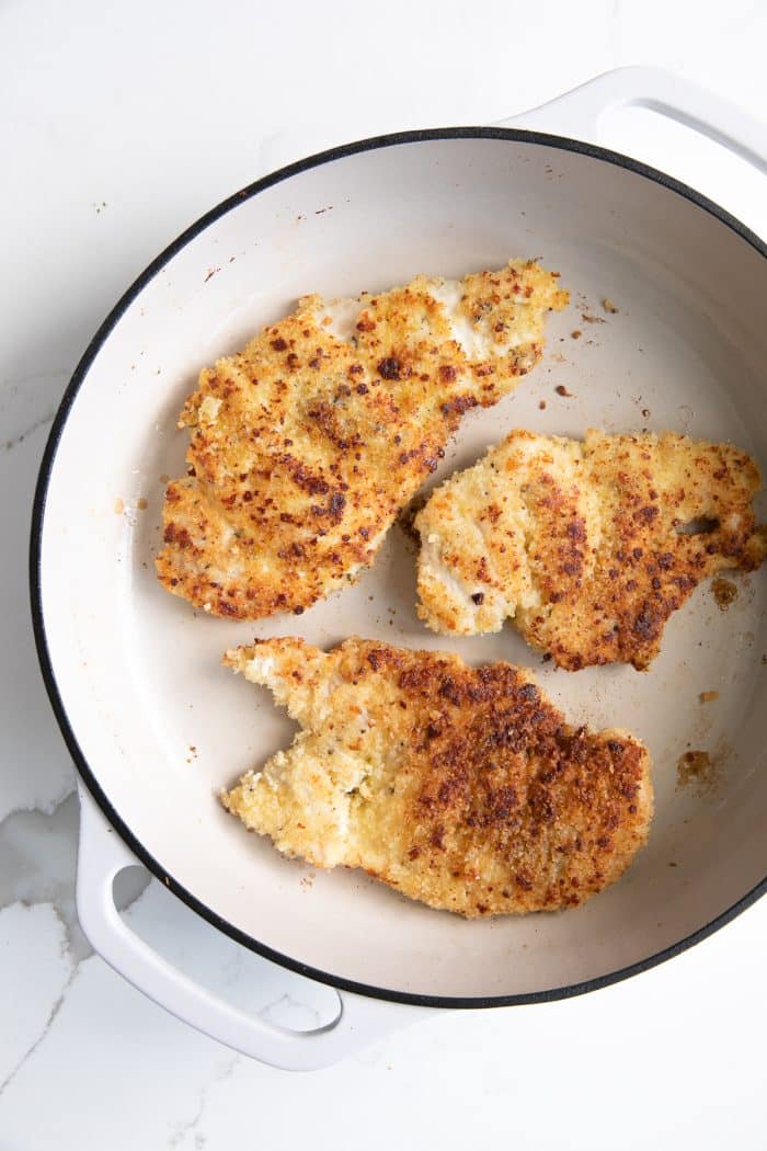 Three golden brown parmesan crusted chicken breasts cooking in a large pan..