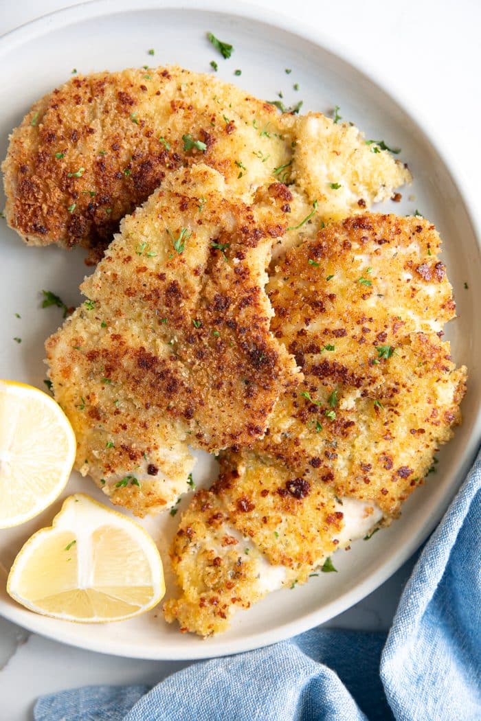 Two thinly sliced parmesan crusted chicken breast pieces on a small white serving plate garnished with chopped parsley and fresh lemon.