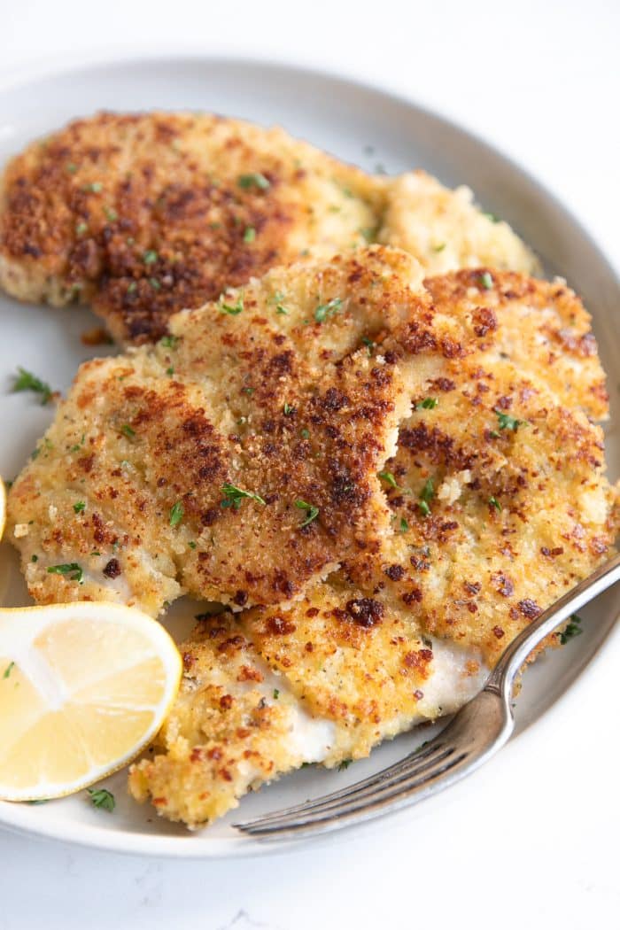 Close up image of thinly sliced parmesan crusted chicken breast pieces on a small white serving plate garnished with chopped parsley and fresh lemon.