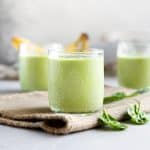 Several glasses of icy cold and creamy homemade green smoothie resting on a folded burlap placemat with fresh bananas in the background and fresh spinach leaves scattered to the side.