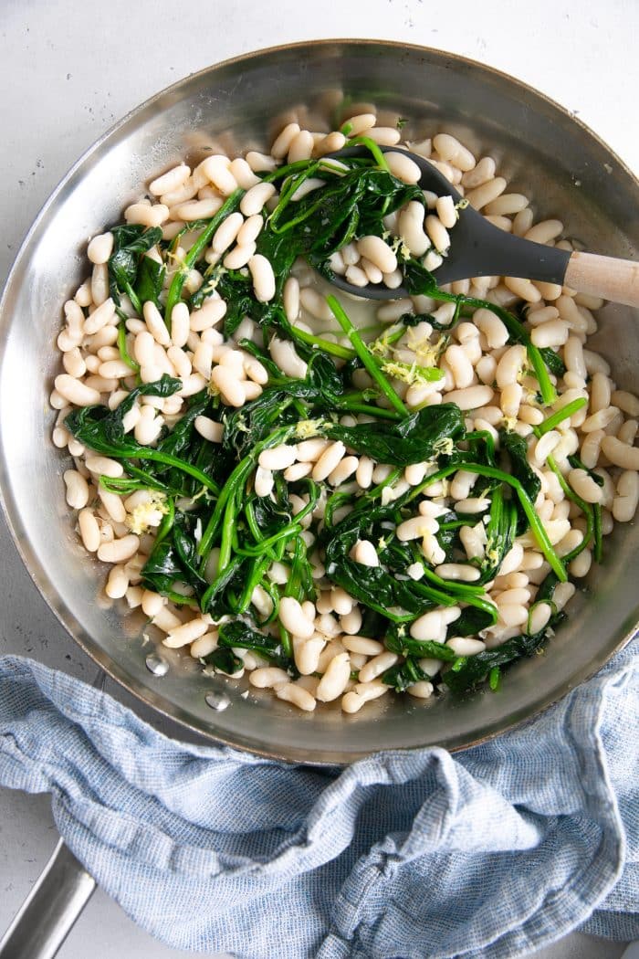Overhead image of a large stainless steel skillet filled with cooked cannellini beans and spinach in a savory lemon and garlic sauce.