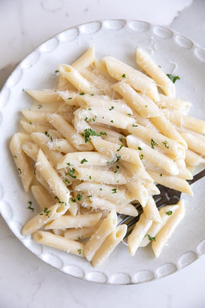 Small white plate with penne noodles tossed in a homemade cream sauce and garnished with fresh parmesan and lemon zest.