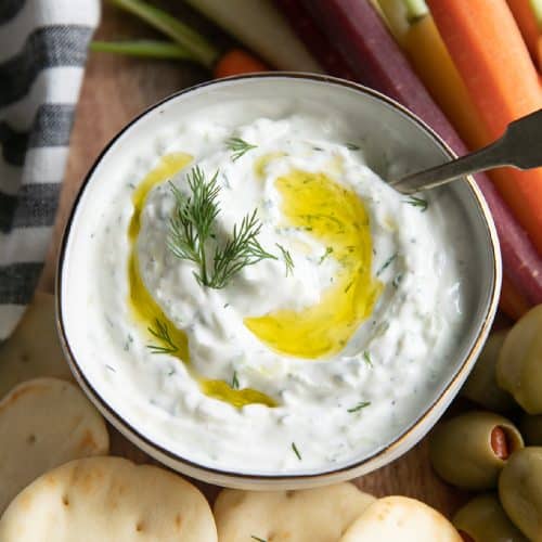 Small white serving bowl filled with homemade tzatziki sauce recipe drizzled with olive oil and garnished with fresh dill.