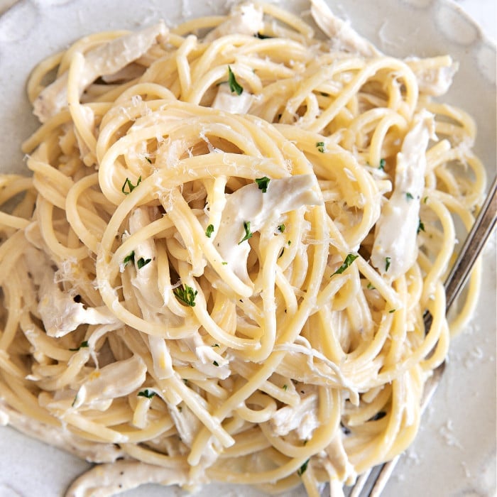 White plate filled with spaghetti noodles and shredded chicken coated in a lemon creamy sauce and garnished with parmesan cheese and chopped parsley.