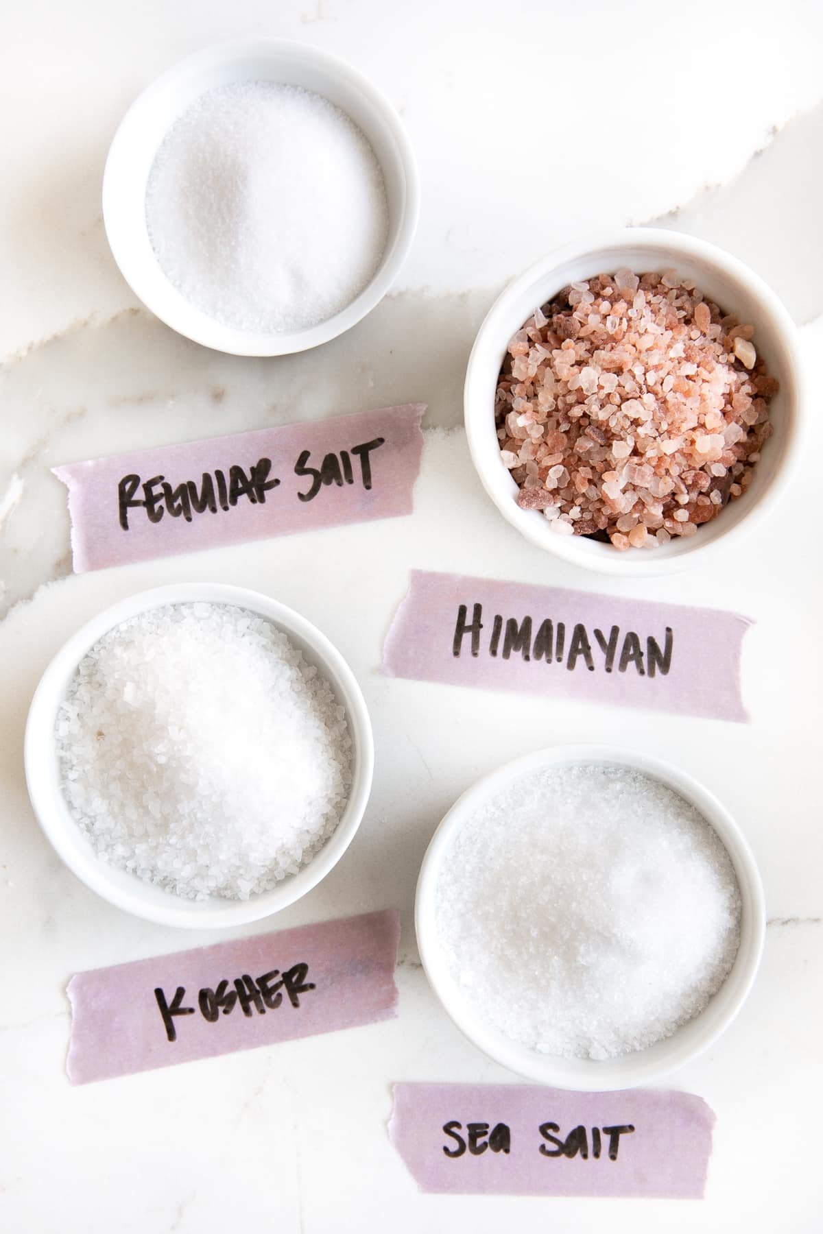 Image of the four types of salt (regular salt, Himalayan, kosher, and sea salt) each in their own white ramekin and labeled on light purple tape.