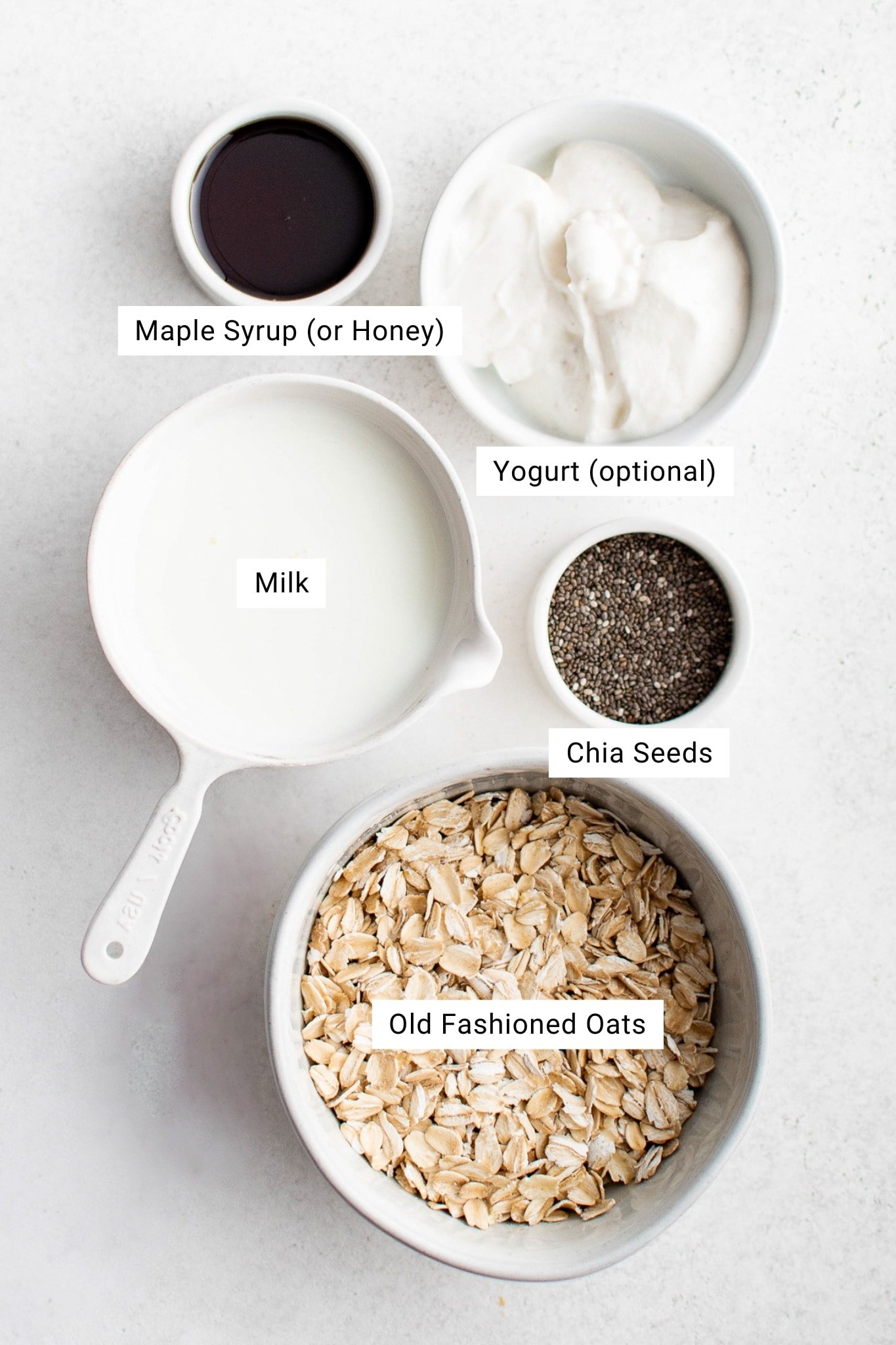 Overhead image of labeled ingredients for overnight oats, including a bowl of old-fashioned oats, milk, yogurt, chia seeds, and maple syrup or honey, each placed in a separate container on a white background.