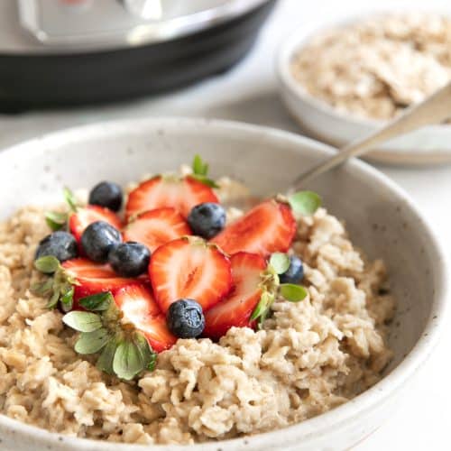 Image of a bowl filled with pressure cooker oatmeal topped with fresh strawberries and blueberries with a pressure cooker in the background.