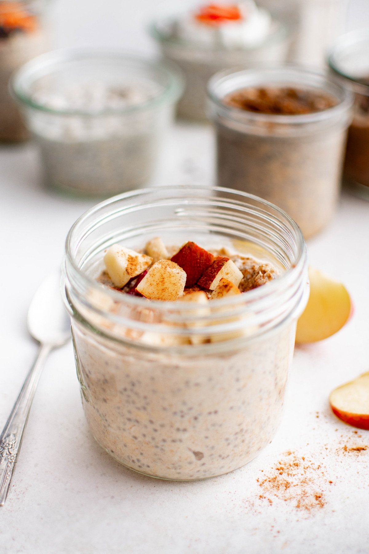 A jar of apple pie overnight oats topped with chopped apples and sprinkled with cinnamon. A spoon rests beside the jar with slices nearby.
