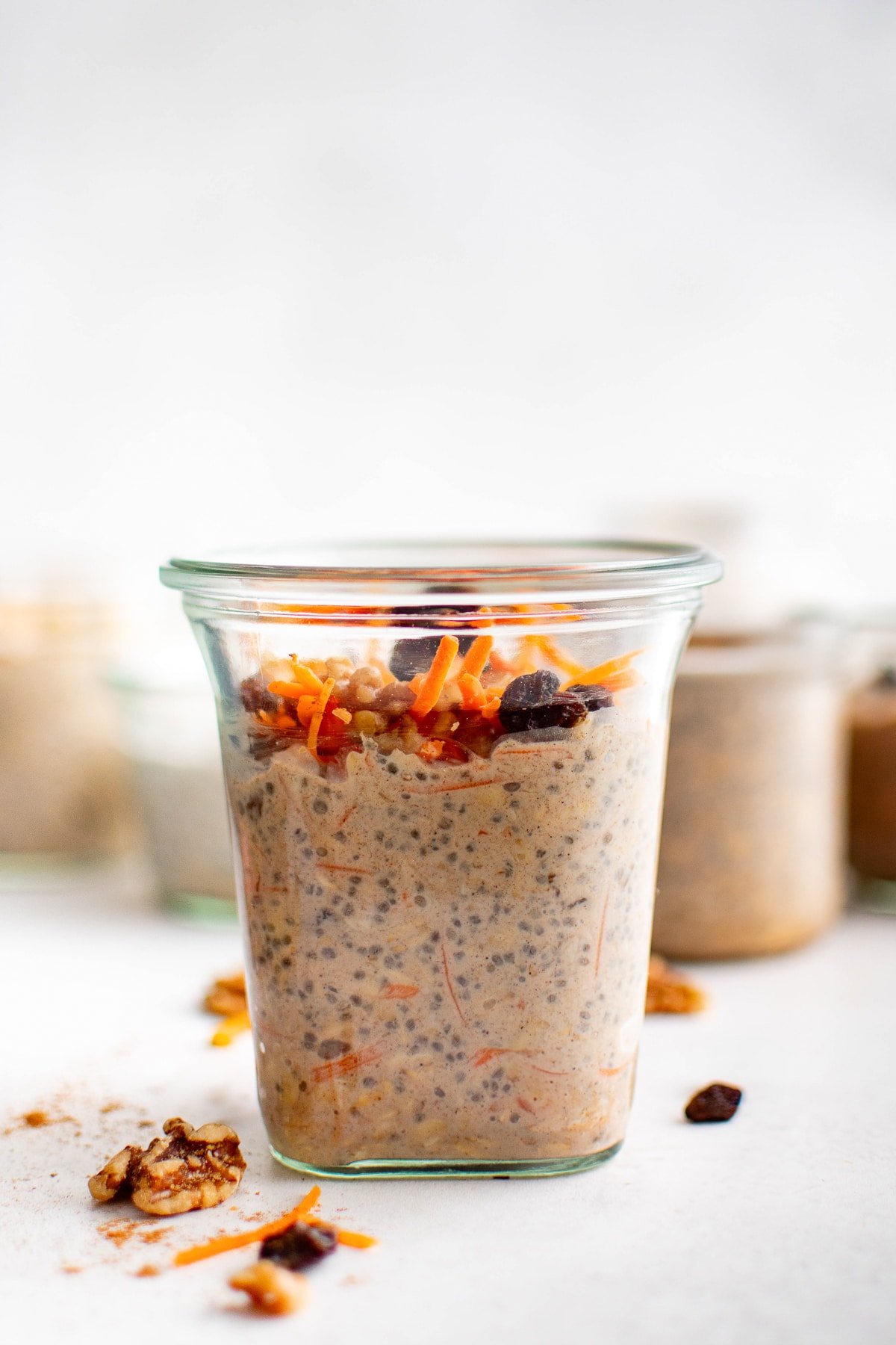 Tall glass jar of carrot cake overnight oats with grated carrots, raisins, and chopped walnuts on top. Background jars are softly blurred.