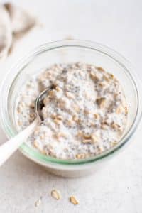 Overhead close-up of stirred overnight oats with chia seeds in a glass jar. A spoon rests inside the jar, showing the thick and creamy texture.