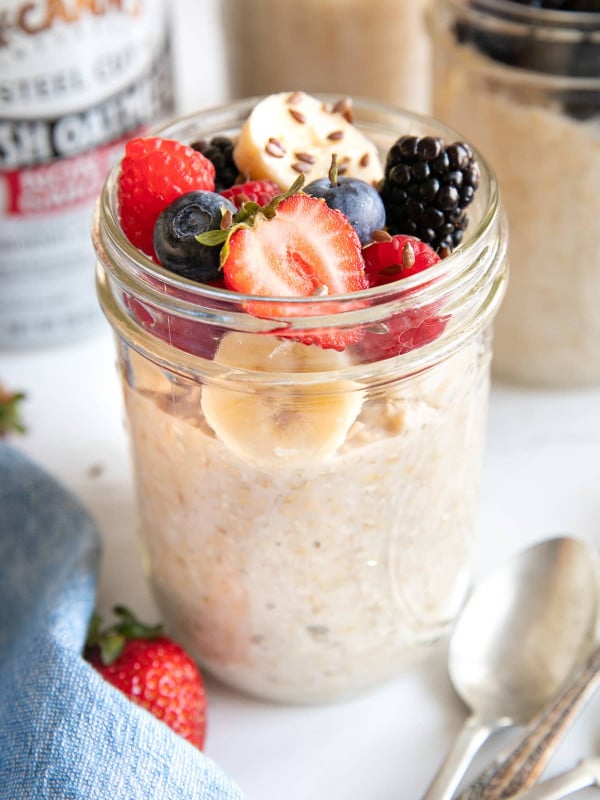 Glass mason jar filled with overnight steel cut oats and topped with sliced banana, strawberries, blueberries, raspberries, and blackberries, and sprinkled with flax seeds.