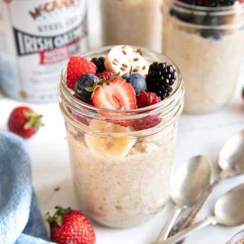 Glass mason jar filled with overnight steel cut oats and topped with sliced banana, strawberries, blueberries, raspberries, and blackberries, and sprinkled with flax seeds.