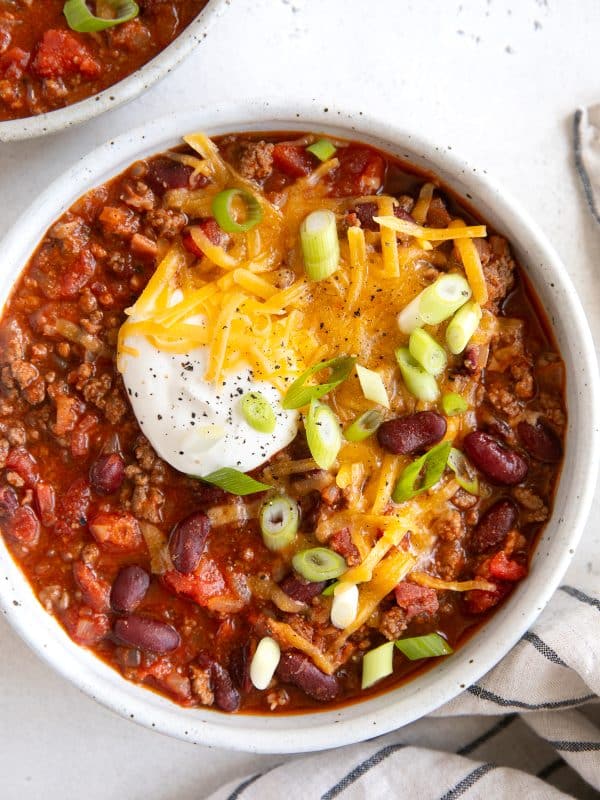 White ceramic bowl filled with chili topped with shredded cheddar cheese, a dollop of sour cream, and sliced green onions.