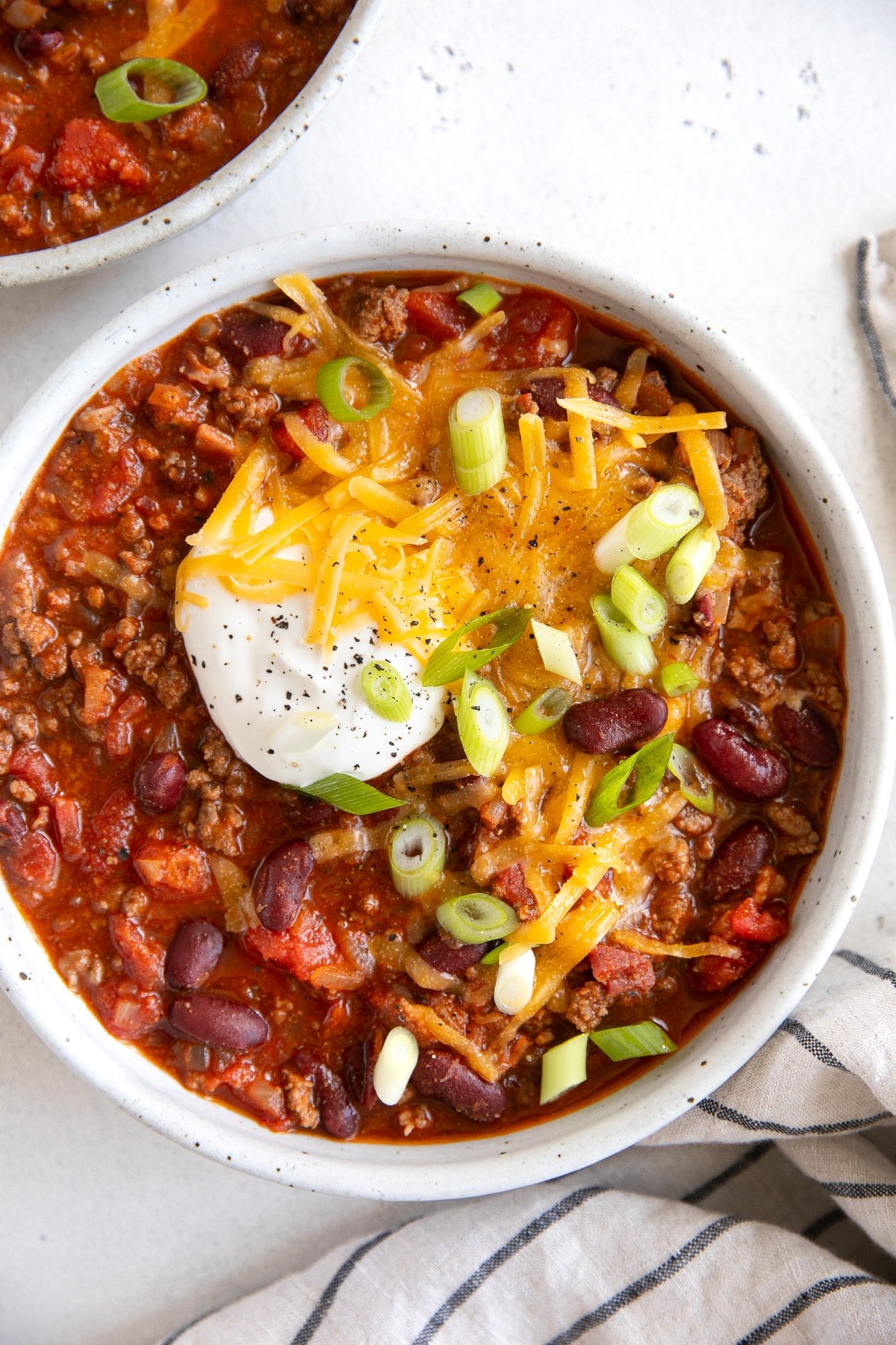 White ceramic bowl filled with chili topped with shredded cheddar cheese, a dollop of sour cream, and sliced green onions.