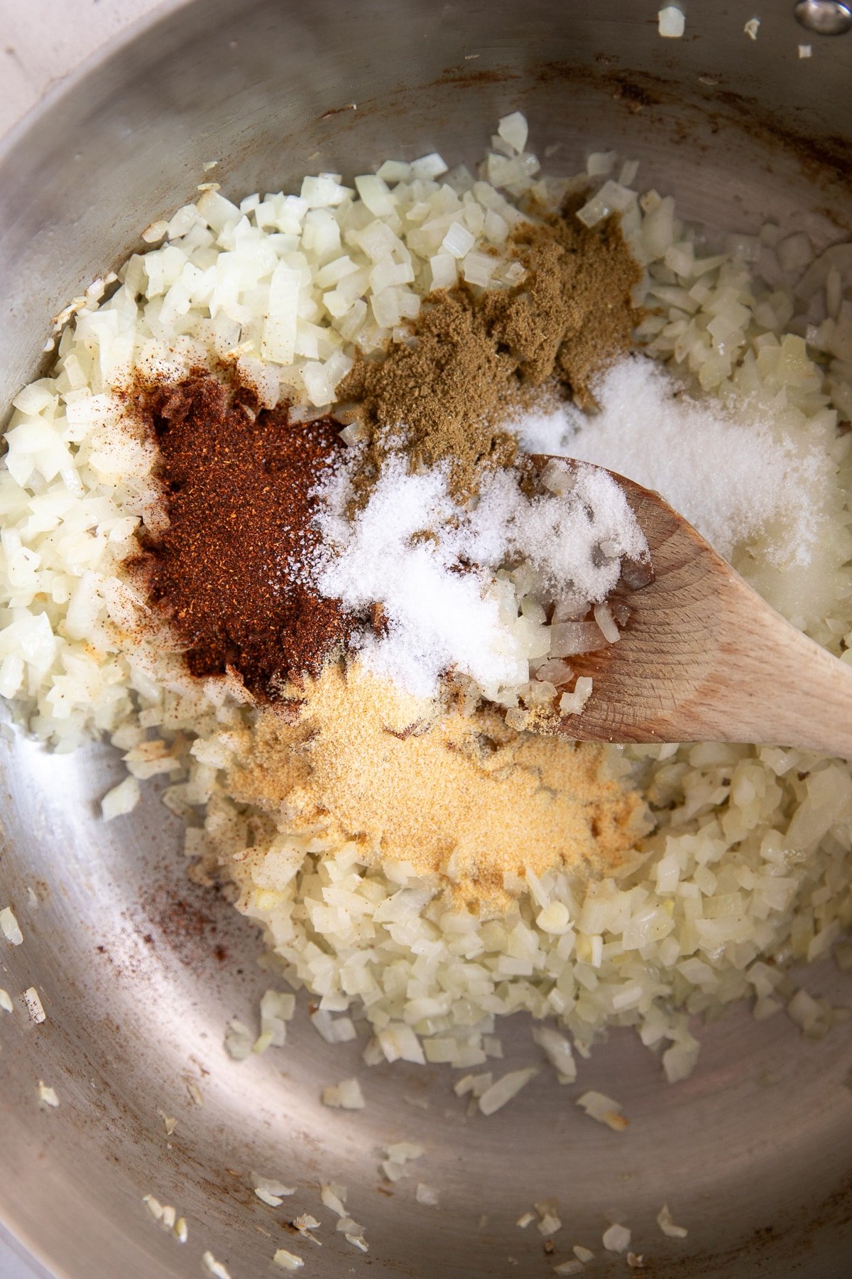 Chopped white onions cooking in a stainless-steel pot, topped with piles of spices including chili powder, cumin, garlic powder, and salt. A wooden spoon rests on the onions.