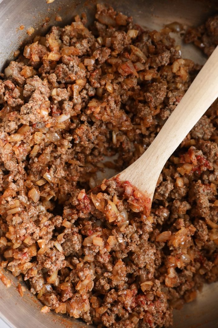 Wooden spoon stirring together browned beef, diced onions, spices, and tomato paste until well combined in a pot.