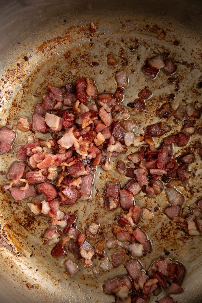 Small pieces of bacon cooking in a stainless-steel pot, browned and sizzling in rendered bacon grease.