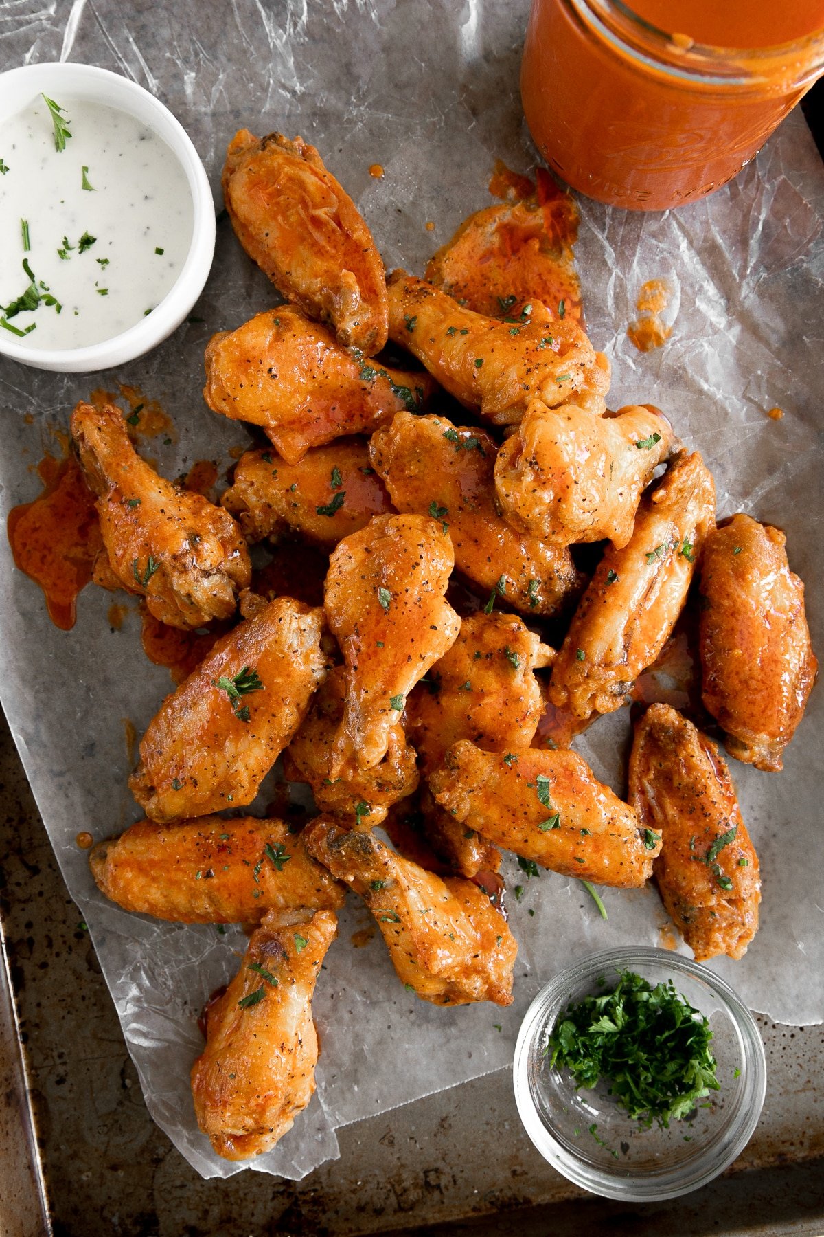Overhead image of a large baking sheet with parchment paper on top piled with buffalo chicken wings served with a side of ranch dressing, fresh chopped parsley, and additional buffalo sauce.