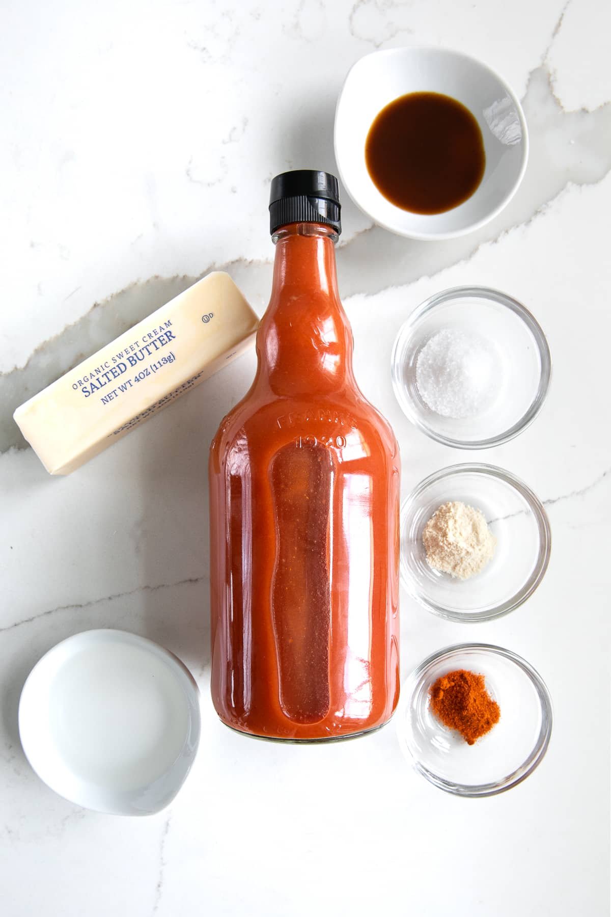 Overhead image of the ingredients needed to make homemade buffalo wing sauce: full bottle of hot sauce, white vinegar, stick of butter, Worcestershire sauce, salt, garlic powder, and cayenne powder.