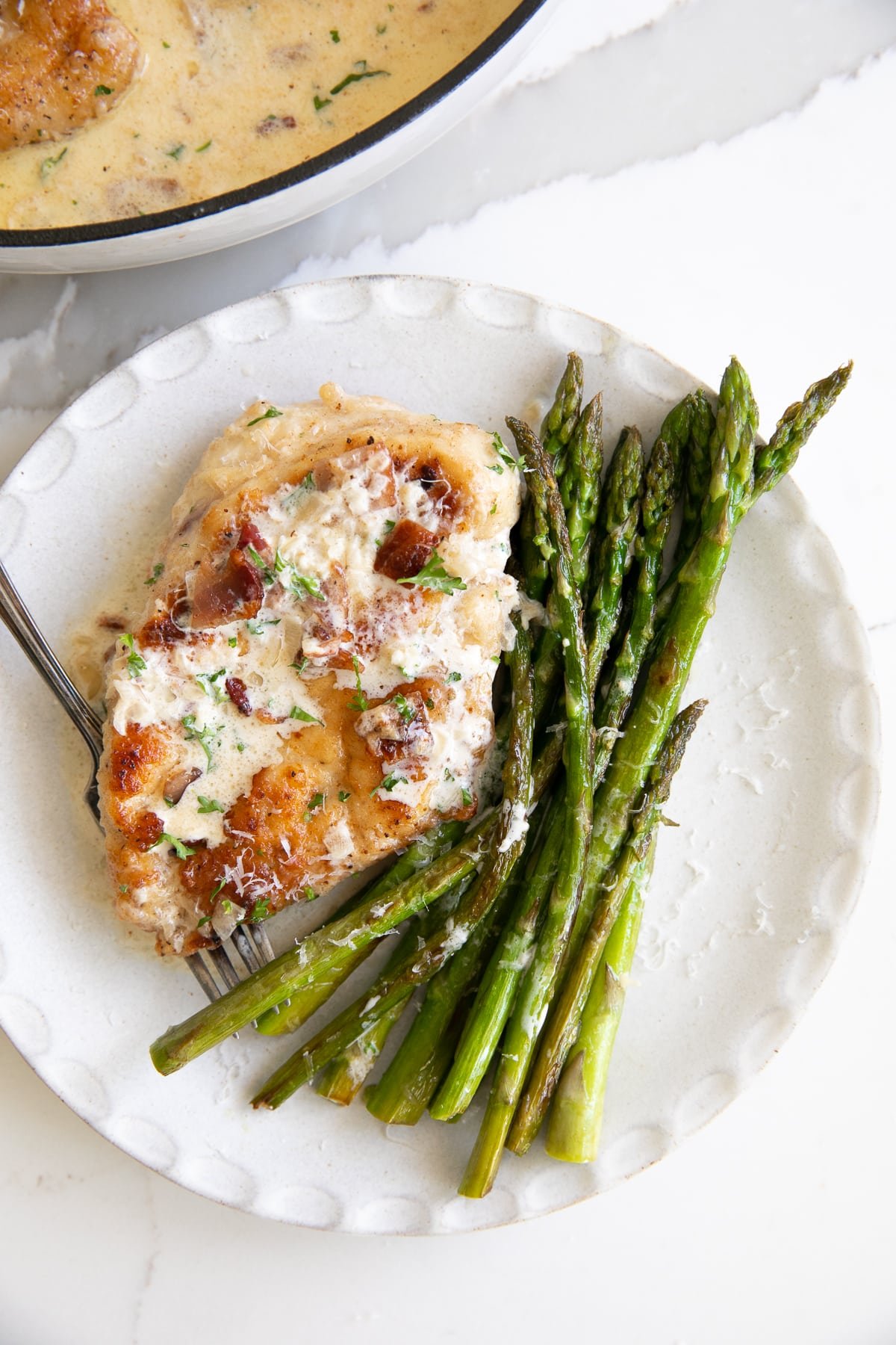 Overhead image of a white plate filled with chicken breast and asparagus drizzled with creamy bacon sauce.
