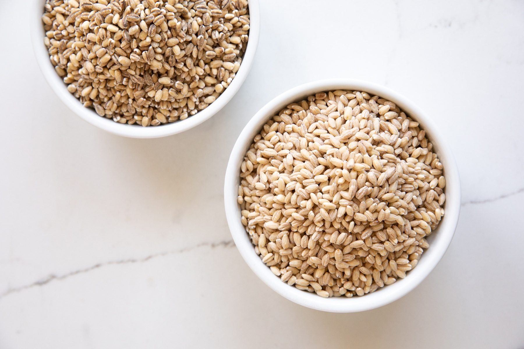 Image of two white ramekins one filled with pearled barley and one with hulled barley.