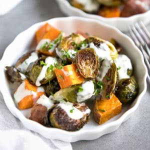 Roasted Brussels sprouts and sweet potatoes topped with a creamy sauce and chopped parsley in a white serving bowl.