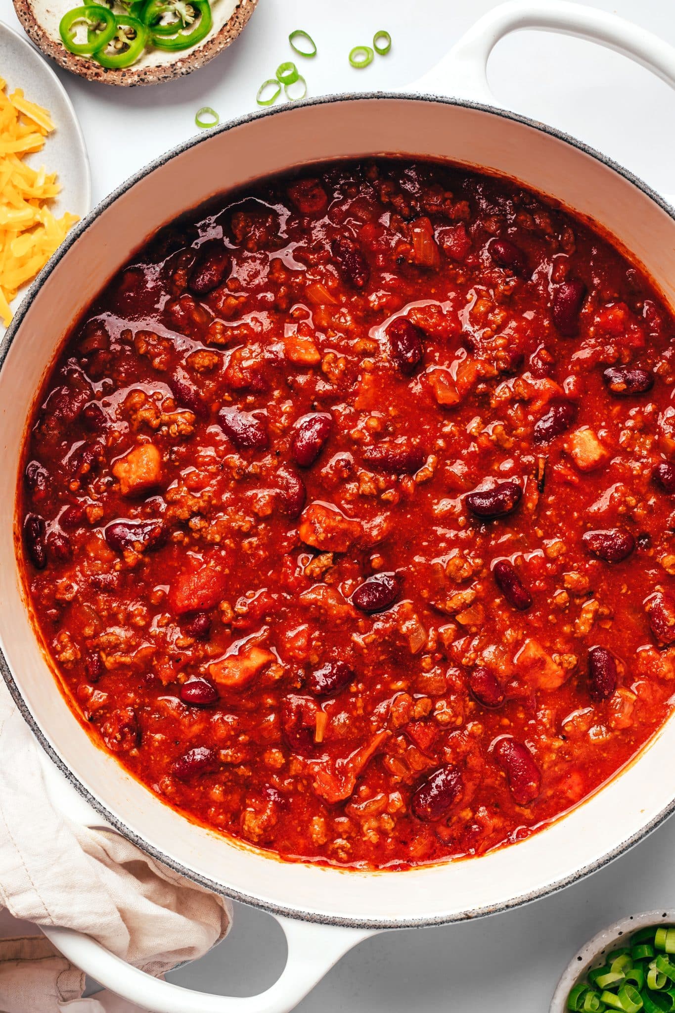 Thick beef and bean chili filling a white Dutch oven, with visible kidney beans, crumbled ground beef, pieces of bacon, and onions in a deep red tomato base.