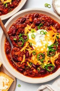 Bowl of beef and bean chili topped with sour cream, shredded cheddar cheese, sliced jalapenos, and green onions, with cornbread and additional chili nearby.