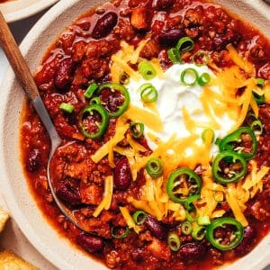 Bowl of beef and bean chili topped with sour cream, shredded cheddar cheese, sliced jalapenos, and green onions, with cornbread and additional chili nearby.