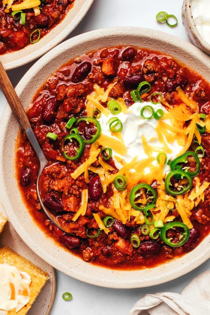 Bowl of beef and bean chili topped with sour cream, shredded cheddar cheese, sliced jalapenos, and green onions, with cornbread and additional chili nearby.