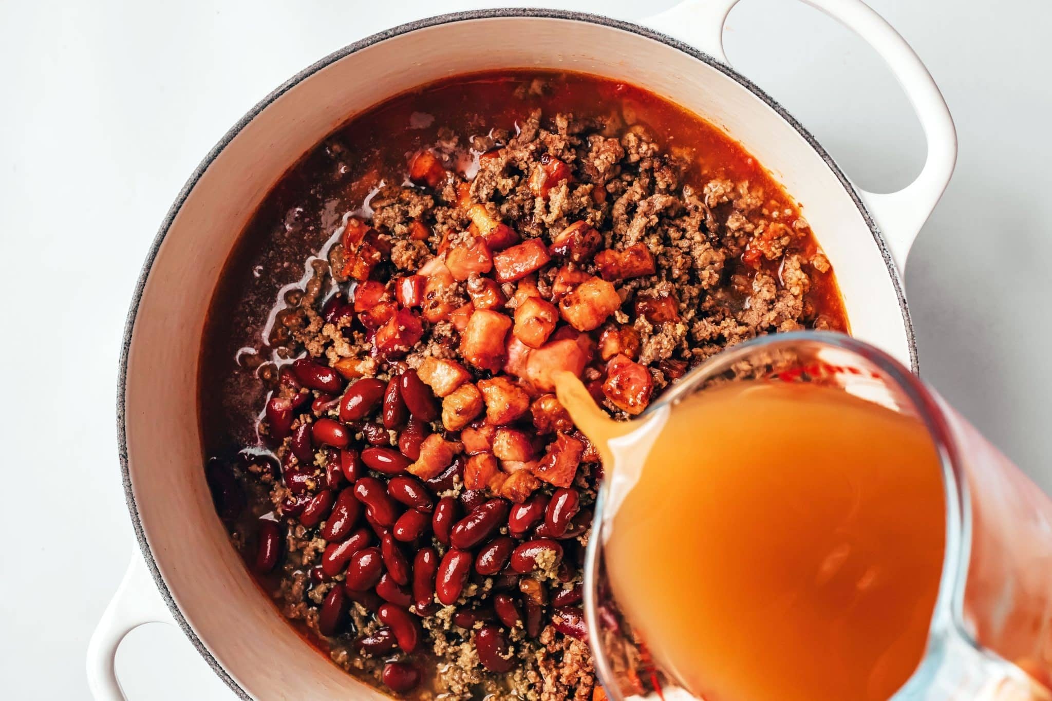 Low-sodium broth being poured from a glass measuring cup into a white Dutch oven filled with ground beef, bacon, kidney beans, and tomato sauce.