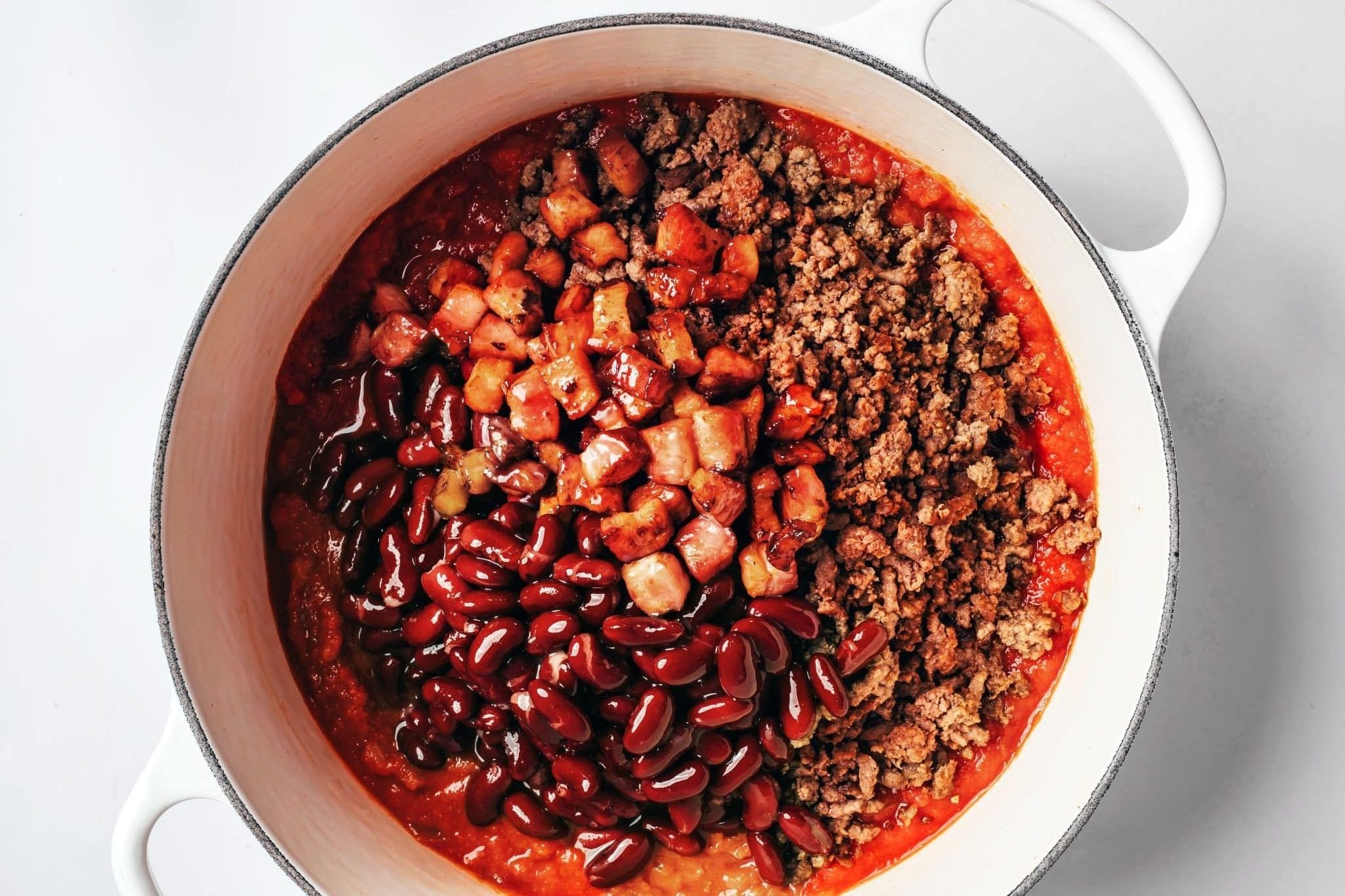 Browned ground beef, crispy bacon pieces, and red kidney beans added to a white Dutch oven over the seasoned tomato and onion mixture.