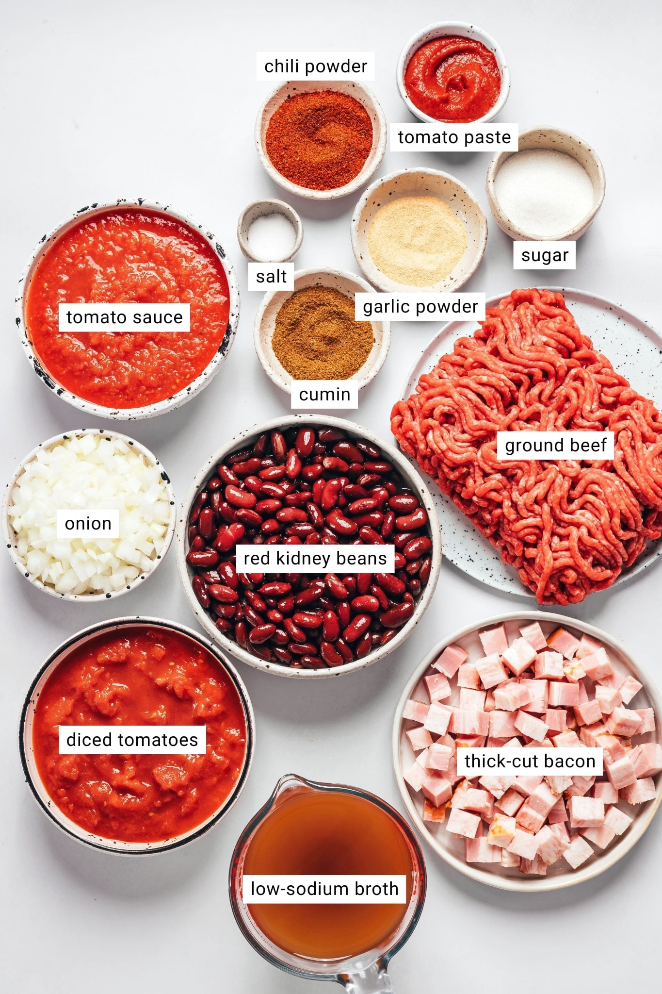 Ingredients for homemade chili arranged in bowls and dishes, including ground beef, thick-cut bacon, kidney beans, diced onion, diced tomatoes, tomato sauce, broth, tomato paste, chili powder, cumin, garlic powder, sugar, and salt, with each ingredient labeled.
