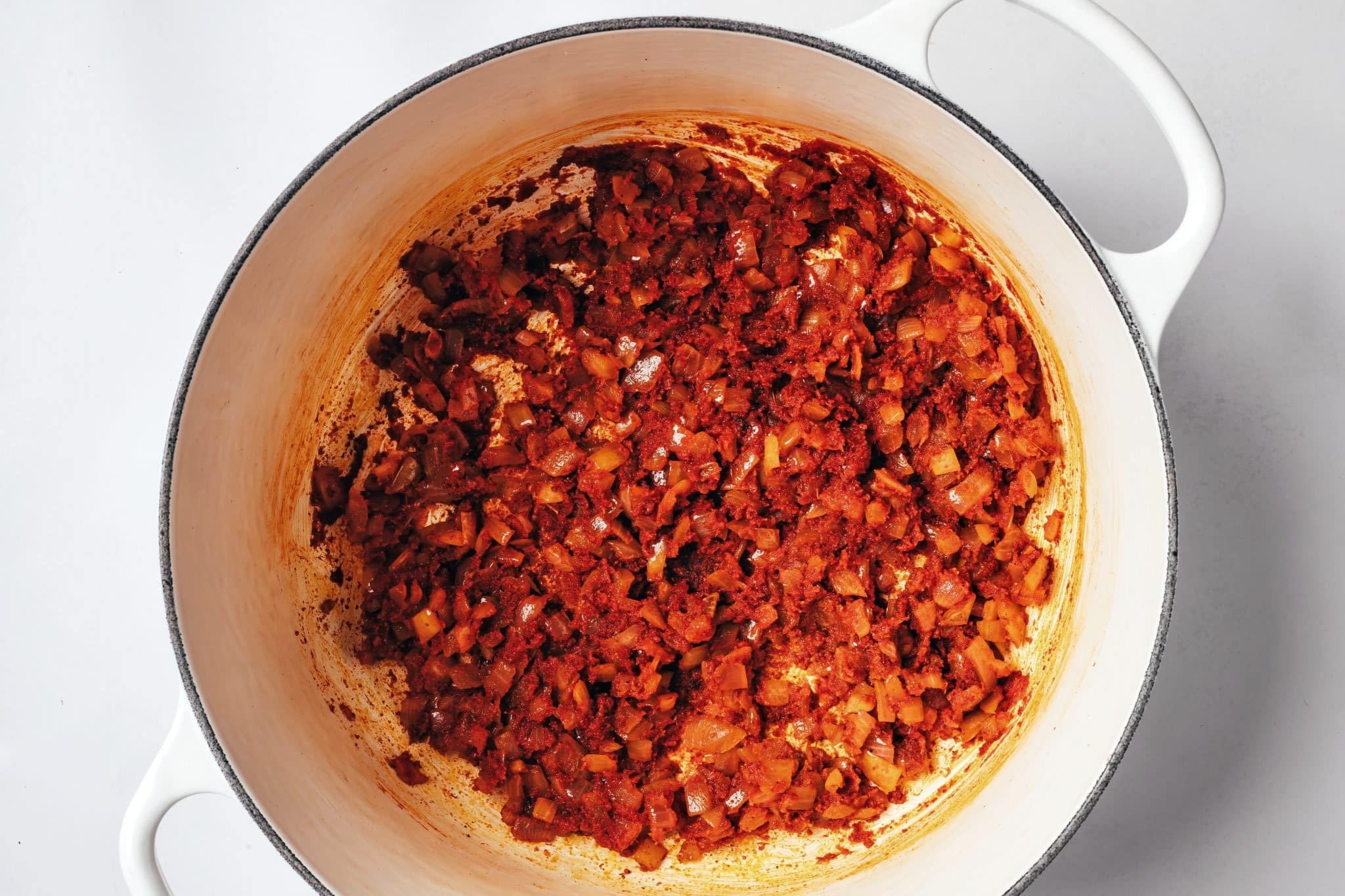 Softened diced onions coated with chili powder, cumin, garlic powder, and tomato paste in a white Dutch oven, forming a deep red, concentrated mixture.