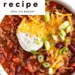 Overhead view of a hearty bowl of beef and bean chili topped with shredded cheddar cheese, sour cream, and sliced green onions. The bowl sits on a white surface, and bold text overlay at the top of the image reads: โSecret ingredient chili recipe โ Hint: itโs bacon!โ