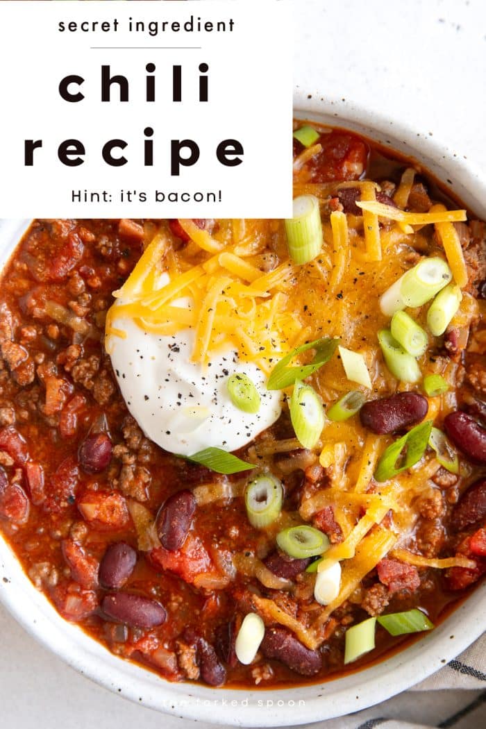 Overhead view of a hearty bowl of beef and bean chili topped with shredded cheddar cheese, sour cream, and sliced green onions. The bowl sits on a white surface, and bold text overlay at the top of the image reads: “Secret ingredient chili recipe — Hint: it’s bacon!”