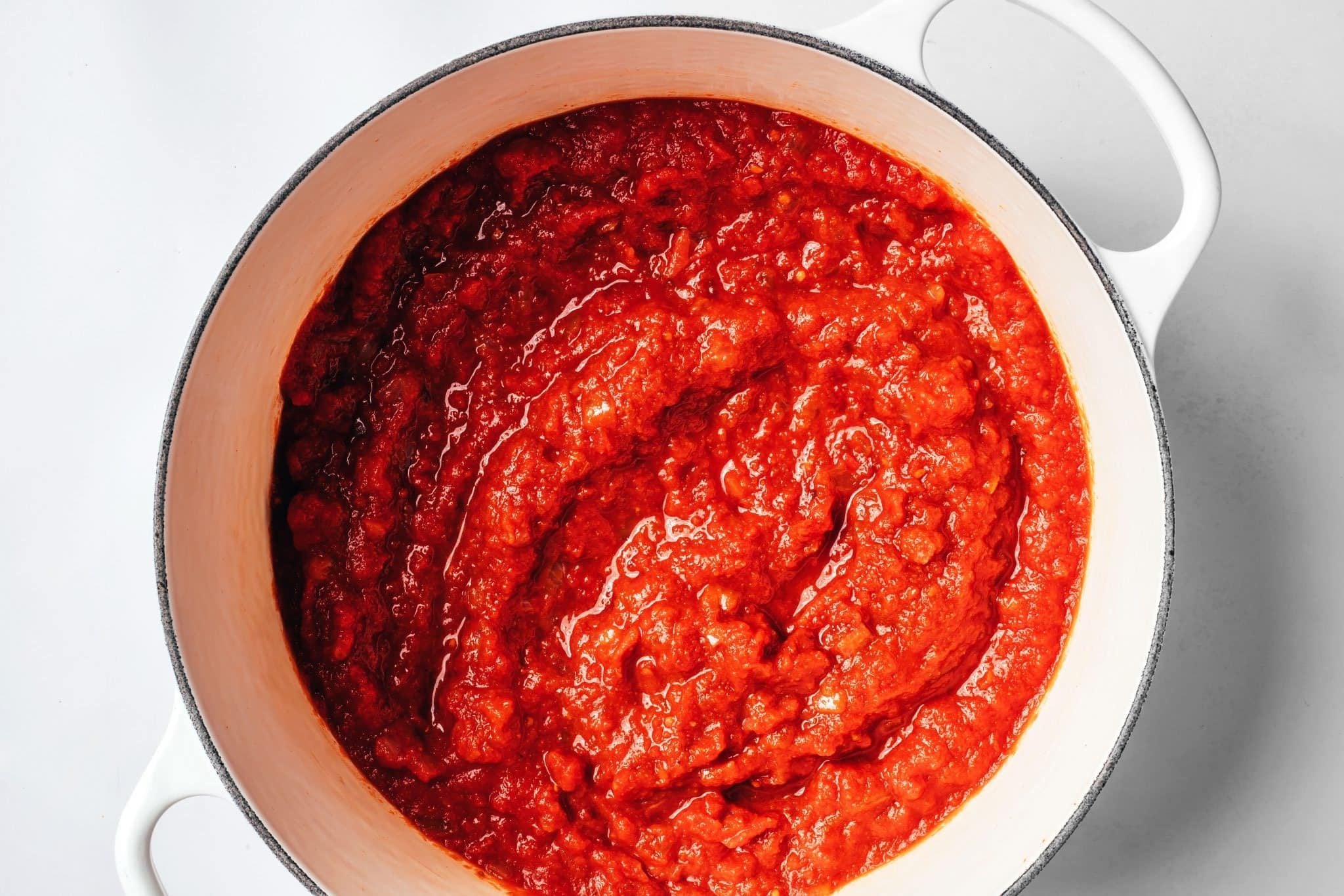 Thick red tomato mixture simmering in a white Dutch oven after the tomato ingredients have been added and stirred together.
