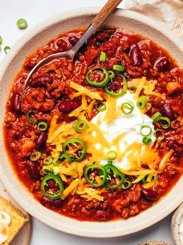 Bowl of thick homemade chili filled with ground beef, kidney beans, bacon, and tomatoes, topped with sour cream, shredded cheddar, sliced jalapenos, and green onions.