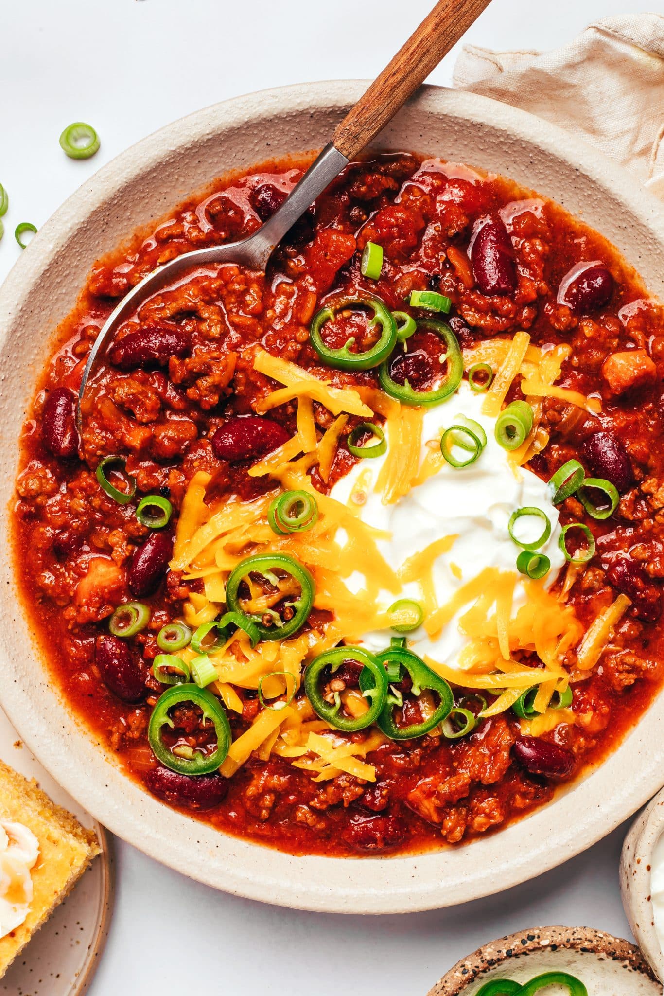 Bowl of thick homemade chili filled with ground beef, kidney beans, bacon, and tomatoes, topped with sour cream, shredded cheddar, sliced jalapenos, and green onions.