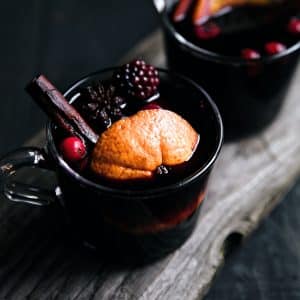 Two glasses filled with warm mulled wine garnished with a cinnamon stick, orange slice, fresh black berries, and cranberry.