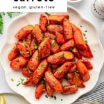 Vertical Pinterest image featuring a scalloped white plate piled high with roasted carrots sprinkled with fresh parsley, surrounded by lemon wedges and a striped napkin, with a white banner at the top that reads โquick & easy roasted carrots, vegan, gluten-free.โ