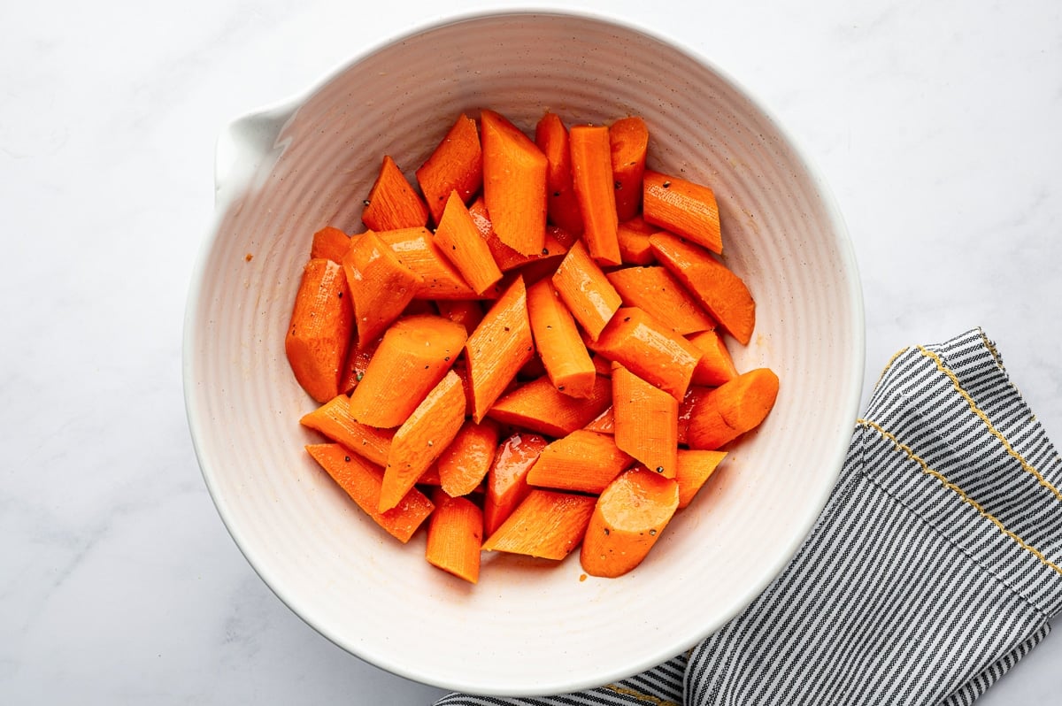 Carrot pieces tossed with oil, salt, and pepper in a white mixing bowl.