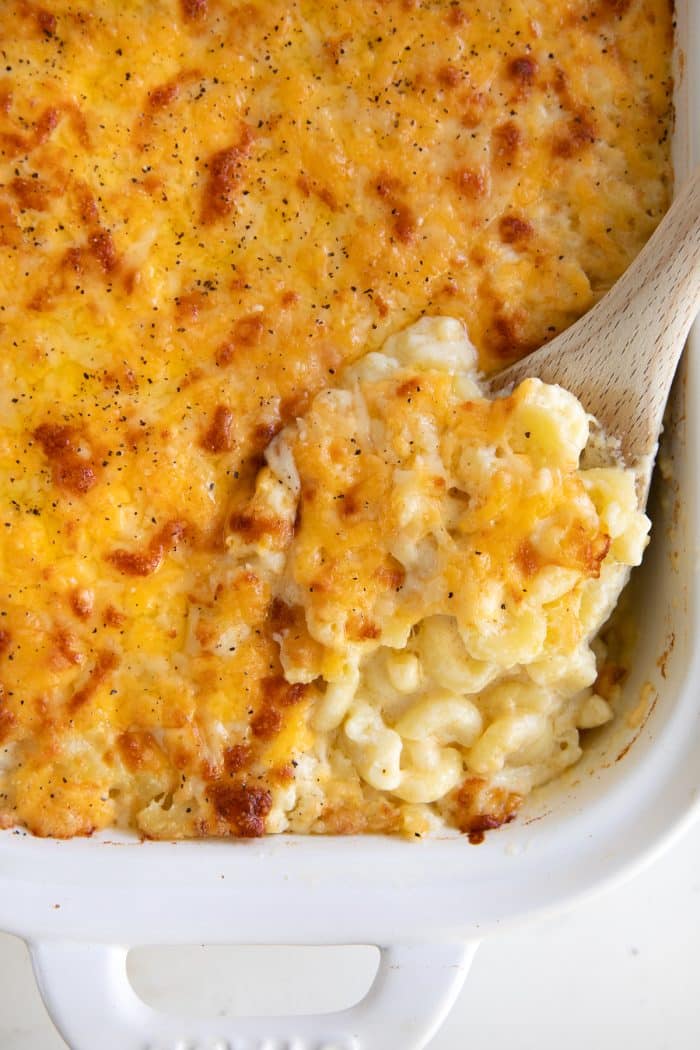 Wooden spoon filled with creamy baked macaroni and cheese in a white baking dish.