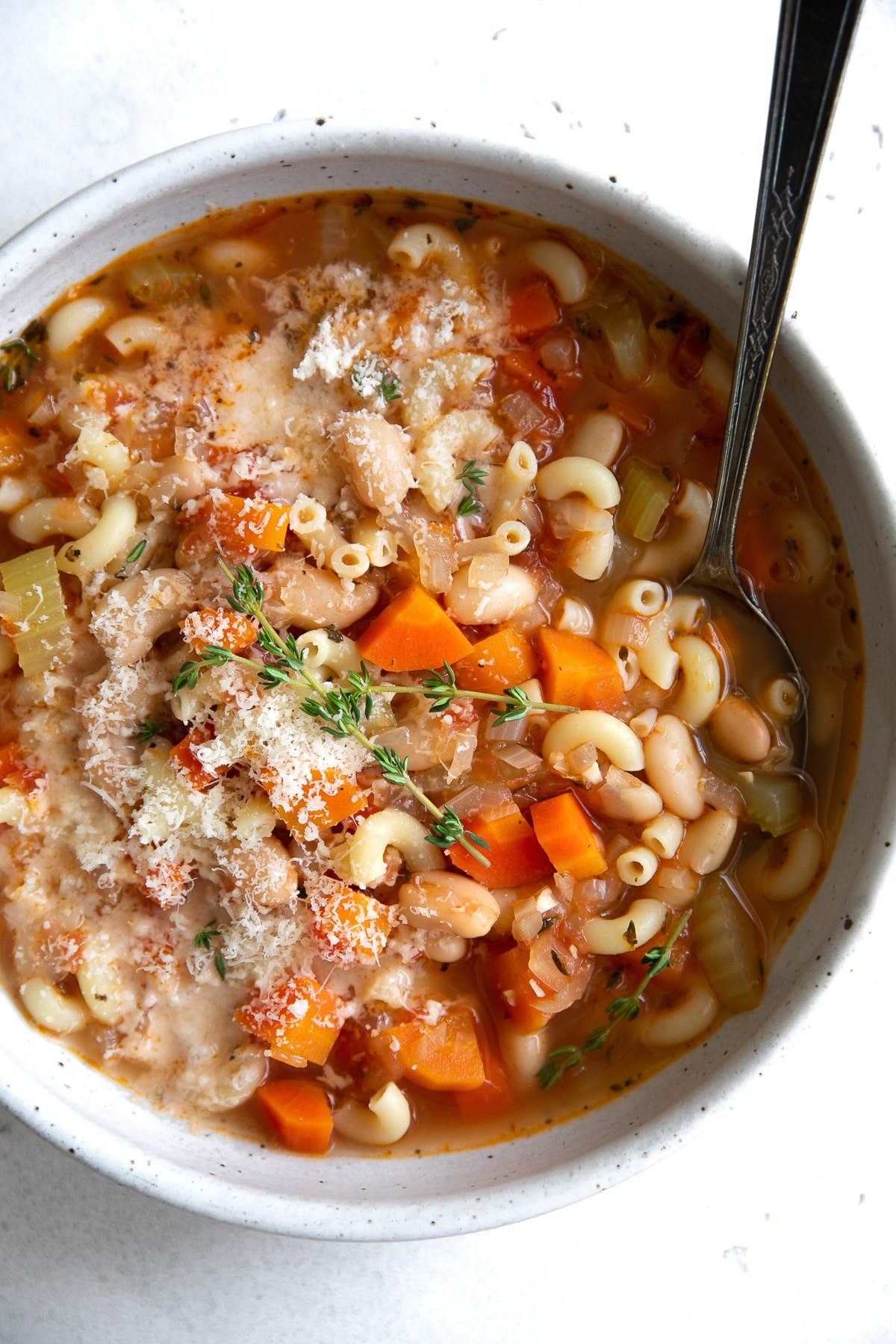 Top down image of a white shallow bowl filled with macaroni pasta, white beans, tomatoes, vegetables, in a light broth.