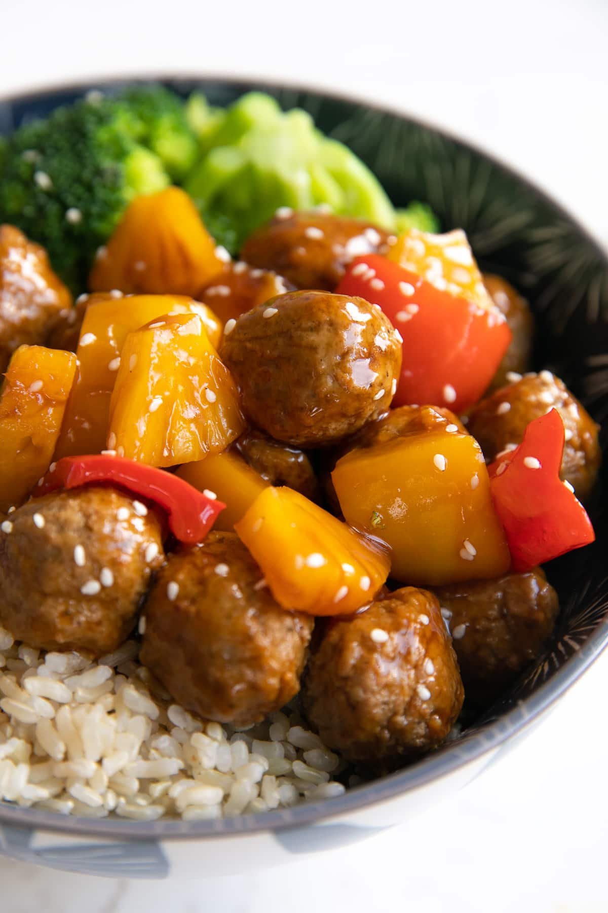 One bowl filled with brown rice topped with sweet and sour meatballs and broccoli sprinkled with white sesame seeds.